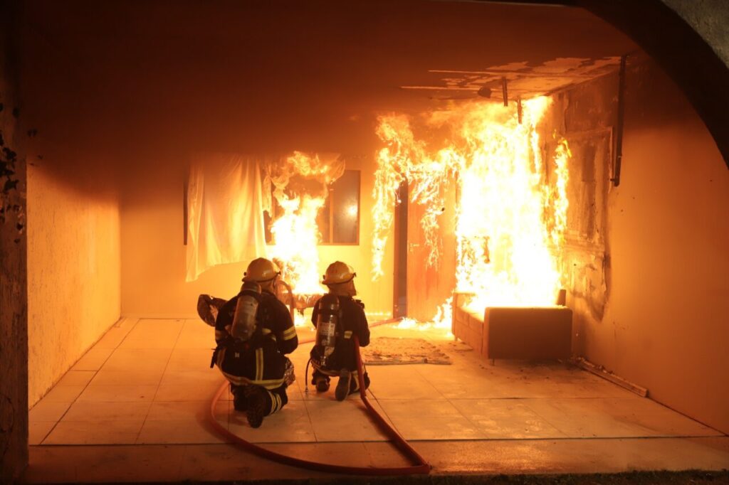 Foto horizontal mostra uma sala pegando fogo e dois bombeiros de costas e de frente para o fogo