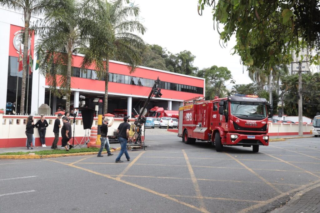 Foto horizontal mostra várias pessoas do lado de fora do prédio do Corpo de Bombeiros Voluntários de Joinville. Há equipamentos de filmagem que acompanham um caminhão vermelho do Bombeiros saindo do estacionamento. O prédio é grande e tem a fachada vermelha.