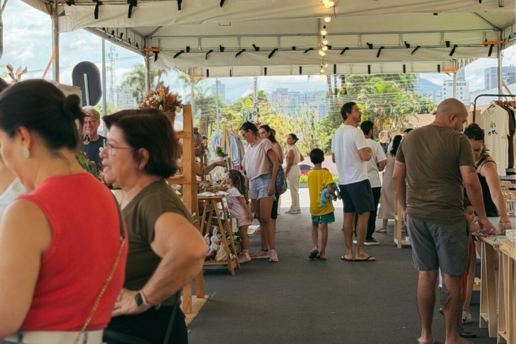 Foto horizontal mostra 15 pessoas, de diferentes idades e características olhando os expositores participantes do Festival Meraki na Praça Cidade das Águas. Os expositores estão embaixo de uma grande tenda branca. Ao fundo e desfocados aparecem prédios, árvorese o céu azul. 
