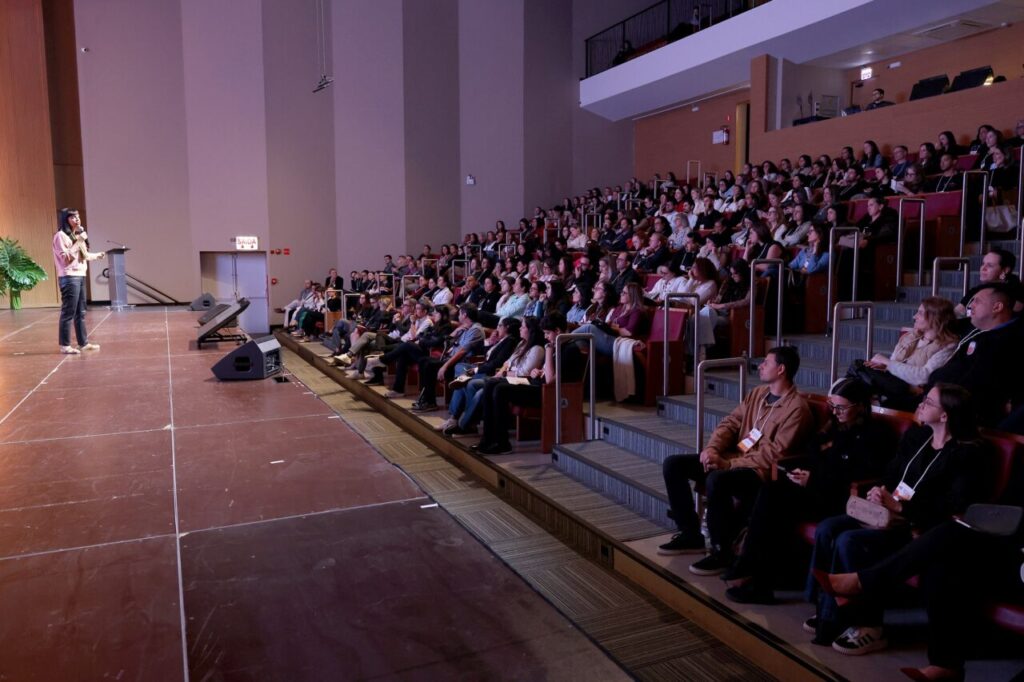 Foto horizontal mostra uma perspectiva lateral do teatro da Coree International School. À esquerda, uma mulher de meia-idade e de cabelos pretos fala ao microfone em um grande palco de madeira, durante o Simpósio de Educação Inovadora Coree 2026. À direita, centenas de pessoas sentadas em cadeiras estofadas em uma plateia, disposta em estilo arena, com vários degraus. 