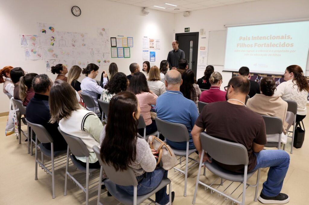 Foto horizontal de um workshop durante o Simpósio de Educação Inovadora Coree 2026. A imagem mostra uma sala de aula com chão, paredes e teto claros. Dezenas de pessoas estão sentadas em cadeiras cinzas. À frente delas, um homem de meia-idade e pele clara está em pé e sorri. Atrás dele, um telão com uma apresentação cujo título é: "Pais intencionais, filhos fortalecidos".