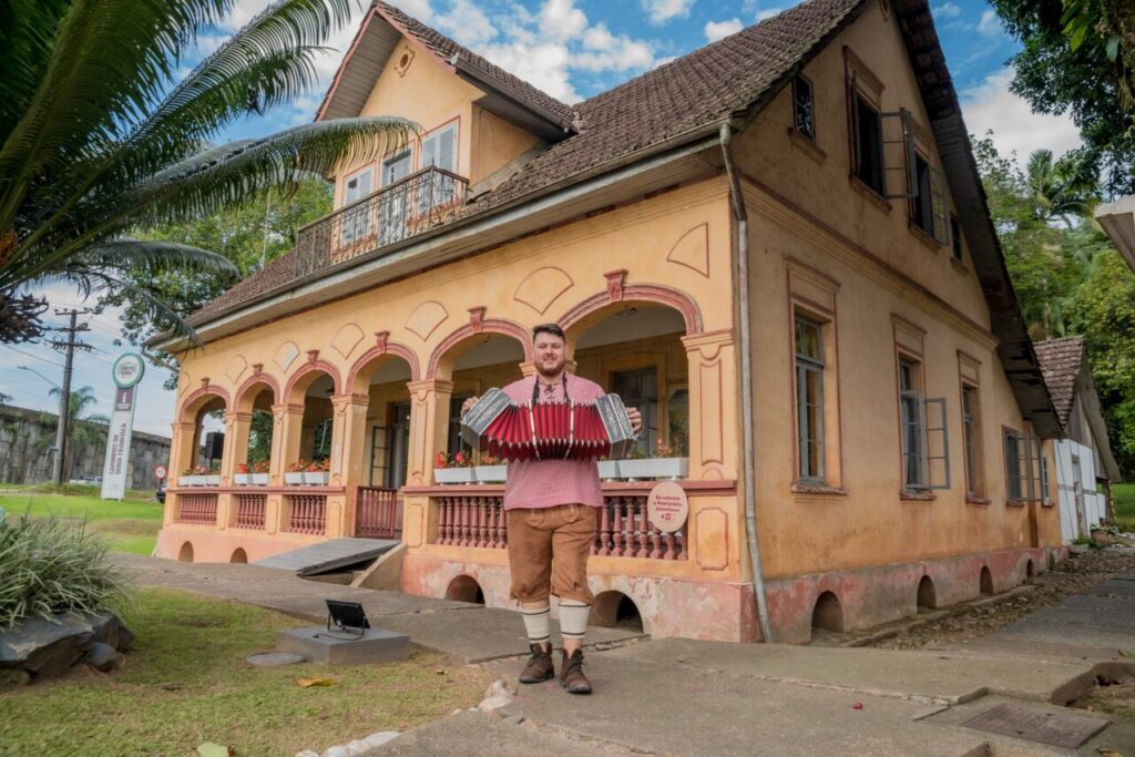 Foto horizontal mostra o músico André Luiz na frente da Casa Krüger. André é um homem jovem, de pele e olhos claros, cabelos curtos loiros e barba cerrada. Ele veste uma camisa quadriculada vermelha, uma calça corcário bege e botas marrons. Ele segura o bandoneon aberto e sorri. Atrás dele está a Casa Krüger, com uma ampla varanda com arcos e balaustres e paredes em tom amarelo envelhecido. Na parte superior da casa há um sótão. O telhado é de duas águas. Ao redor da casa, uma área gramada e árvores.