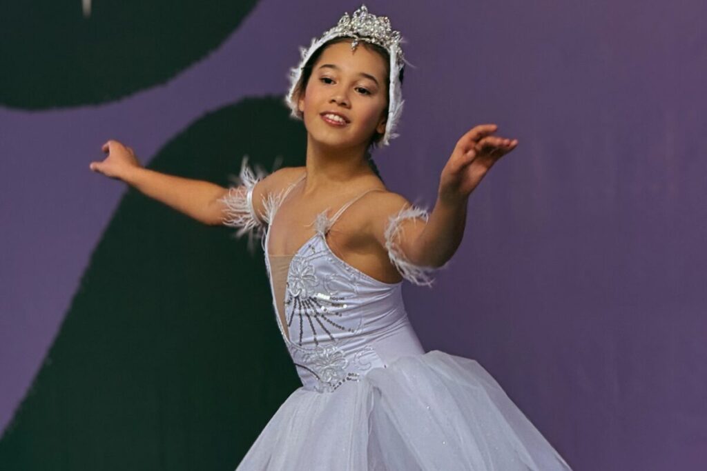 Foto horizontal mostra uma bailarina jovem, de pele parda. Ela usa um vestido com branco, com acessório também brancos na cabeça e nos braços. Ela está com o corpo levemente abaixado, os dois braços abertos e sorri.