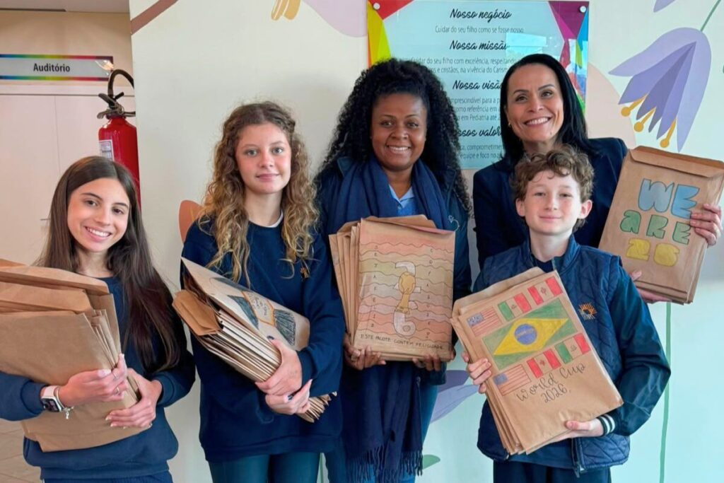 Foto horizontal mostra três estudantes da Coree International School - duas meninas e um menino de peles claras e idade em torno de 12 anos - ao lado de duas mulheres adultas do Hospital Infantil. Uma delas é uma mulher negra e a outra tem pele clara. Todos seguram envelopes pardos com desenhos relacionados à copa do mundo. Eles estão em frente a uma parede colorida do hospital. Olham para foto e sorriem.
