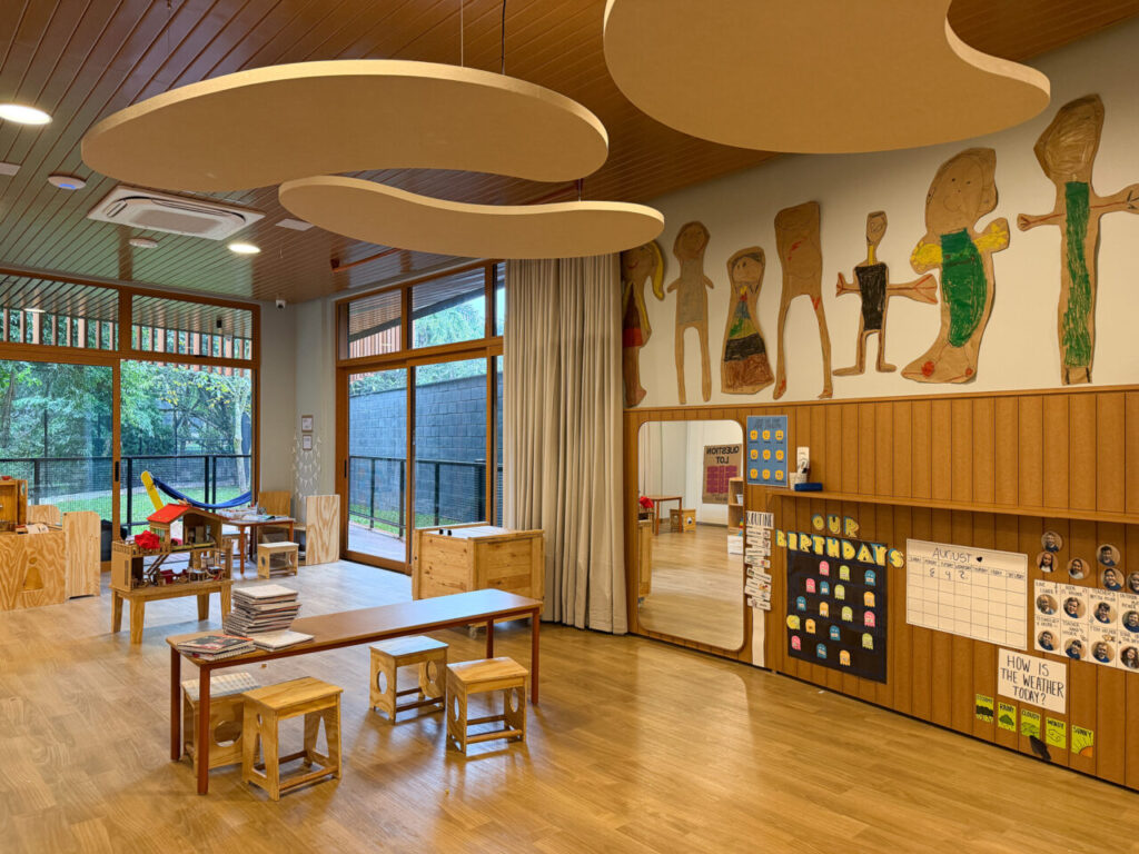 Foto horizontal mostra sala de aula da unidade infantil da Coree International School, com muitos elementos de madeira, grandes portas de vidro e vista para um gramado com árvores.
