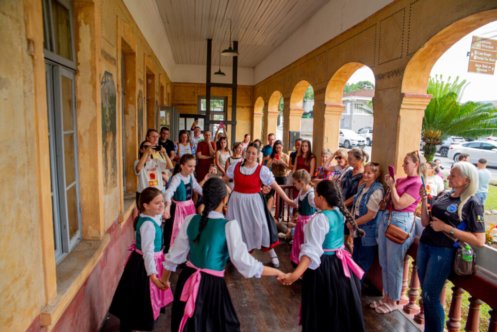 Foto horizontal mostra um grupo de crianças vestidas com trajes germânicos dançando em roda na varanda da Casa Krüger. Ao redor delas dezenas de pessoas assistem à apresentação. 