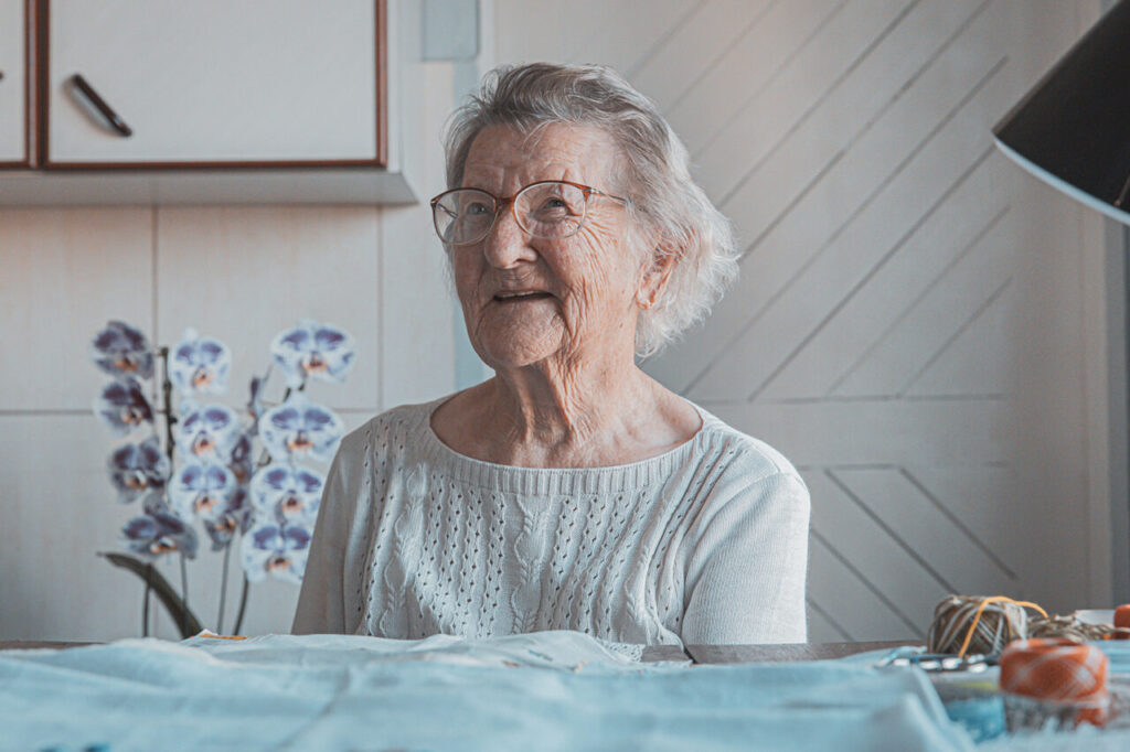 Foto horizontal de dona Ondina Kunde. Ela é uma mulher idosa, de pele e olhos claros, cabelos brancos na altura do queixo. Ela usa um óculos oval claro e veste uma blusa de tricô cinza. Está sentada à frente de uma mesa com tecidos e linhas. Ela olha para o lado e sorri. Ao fundo, um vaso com orquídeas e um armário de cozinha, desfocados.