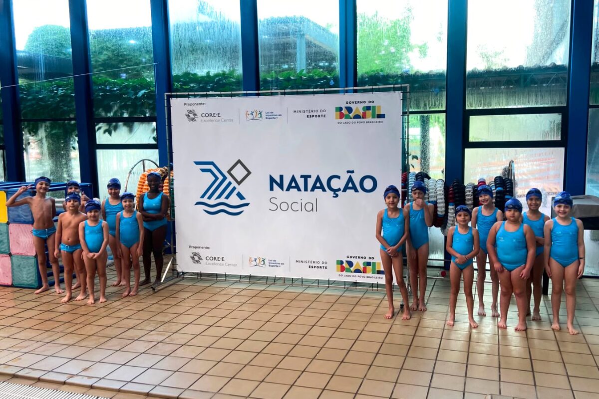 Foto na horizontal mostra 14 crianças de idades entre 7 e 10 anos e diferentes características físicas, dentro do complexo esportivo da Coree International School. Elas usam maiôs e sungas na cor azul claro, toucas em azul escuro e óculos de natação sobre as toucas. Elas estão em pé, divididas em dois grupos, cada um de um lado de um grande painel branco com a logomarca do projeto Natação Social, a logomarca do Instituto Core e dos patrocinadores. O ambiente é coberto e tem grandes paredes de vidro.