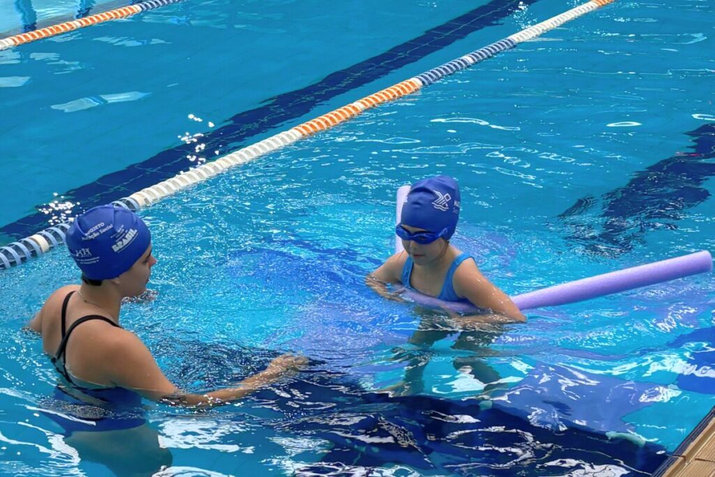Foto na horizontal mostra uma grande piscina com duas raias, no complexo esportivo da Coree International School. Em uma delas há uma menina criança, de pele clara, que usa um maiô na cor azul clara, touca e óculos de natação em azul escuro. Ela está envolvida por uma bóia estilo espaguete. À frente dela, está uma professora de pele clara, que usa um maiô e touca azuis.