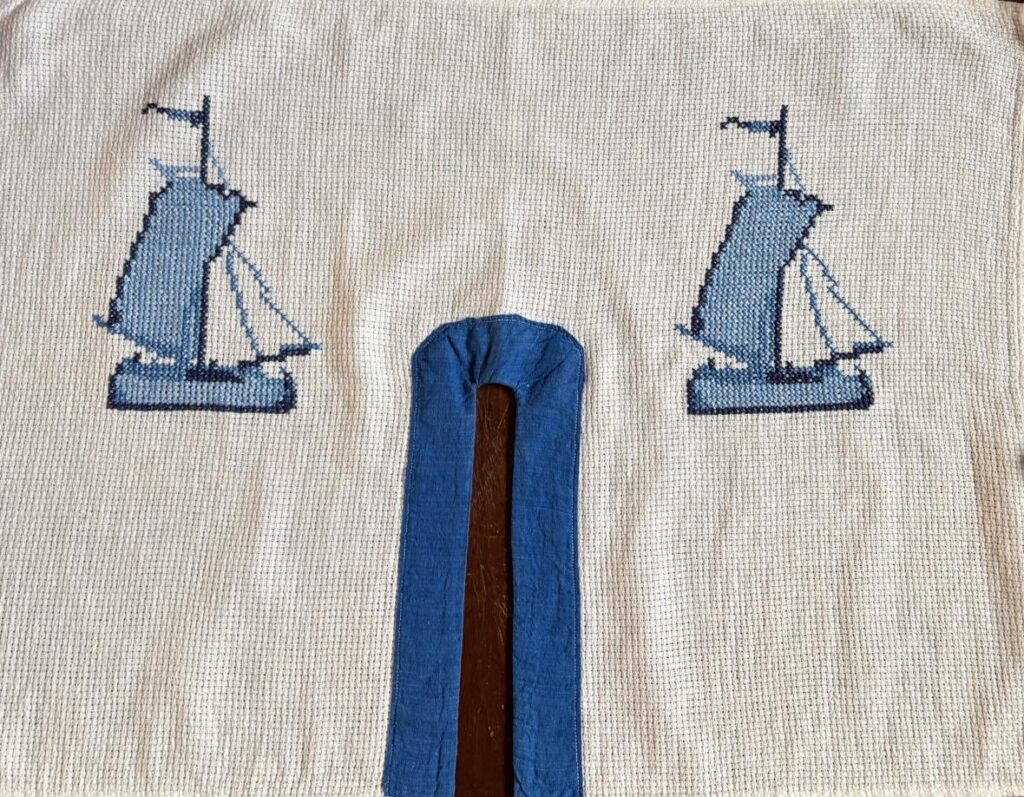 Foto horizontal de um tecido com branco com dois desenhos de um barco à vela, bordado em azul. No meio do tecido há uma fenda, contornada por um tecido azul.