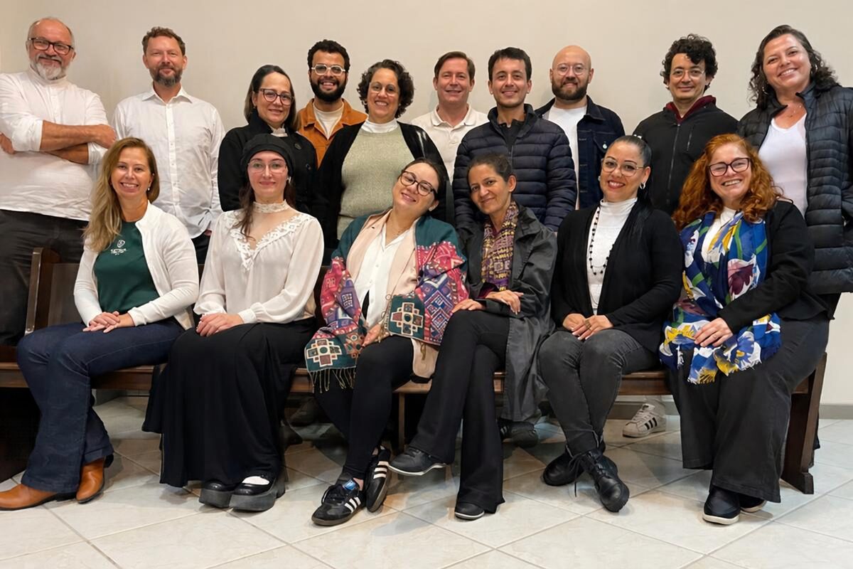 Foto horizontal mostra 16 pessoas entre homens e mulheres de diferentes idades e características físicas. Parte delas está sentada em um grande banco de madeira e outra parte está em pé, atrás do banco. Eles olham para a foto e sorriem. Ao fundo, uma parede clara. 
