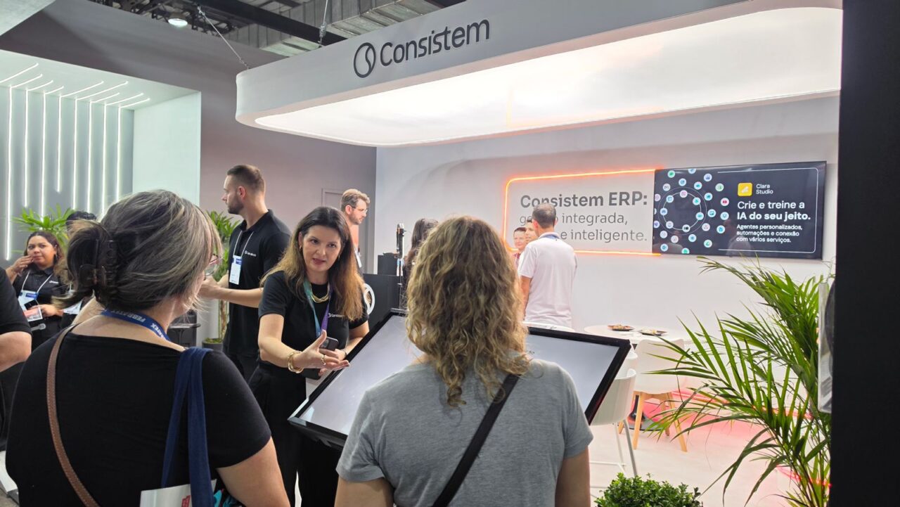 Foto na horizontal mostra o estande da Consistem na Febratex. O espaço tem paredes brancas, mesas e cadeiras claras. Em uma das paredes há um painel com alguns informações, que aparecem parcialmente: "Consistem ERP. Crie e treine a IA do seu jeito". À frente do estande duas mulheres, de costas para foto, conversam com uma mulher de meia-idade e uma blusa preta da Consistem, que gesticula em frente a um totem.