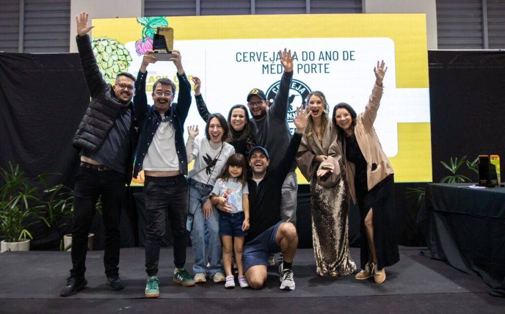 Foto horizontal mostra quatro homens e cinco mulheres, incluindo uma criança, de diferentes idades e características físicas. Eles estão em cima de um pequeo palco, sorriem e estão com as mãos para o alto. Atrás deles, um grande painel, onde aparece parcialmente o escrito: Cervejaria do ano de médio porte e abaixo a logomarca da Stannis, de Jaraguá do Sul.