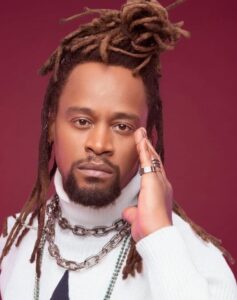 Foto vertical de Toni Garrido. Ele é um homem preto, de meia-idade, rosto alongado, olhos castanhos, lábios grossos, barba e bigode curtos. Seu cabelo tem dreads longos. Ele usa uma blusa de gola alta branca e várias correntes de elo prateadas. O fundo é cor de vinho.