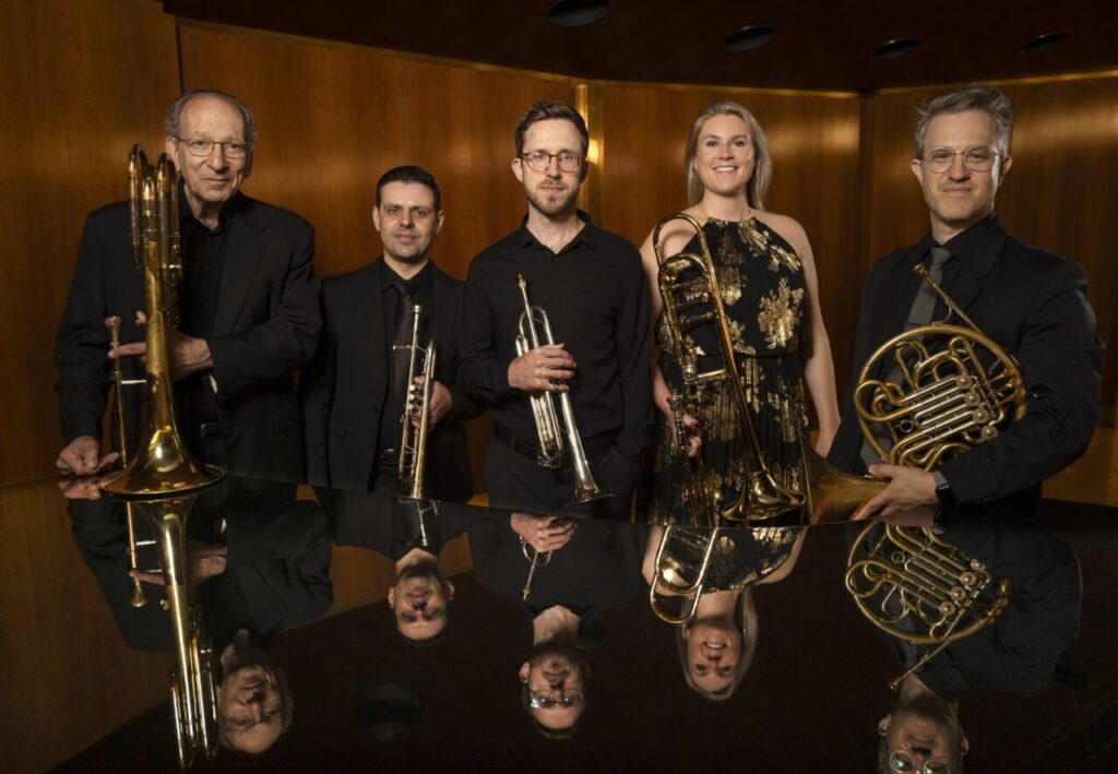Foto na horizontal mostra cinco pessoas lado a lado: quatro homens e uma mulher, integrantes do American Brass Quintet. Todos eles têm pele clara. Eles e vestem roupas sociais pretas, estão em pé, seguram seus instrumentos musicais, olham para foto e sorriem discretamente. À frente deles aparece parcialmente a cauda de um piano, onde a imagem deles é refletida. Ao fundo, uma parede de madeira.