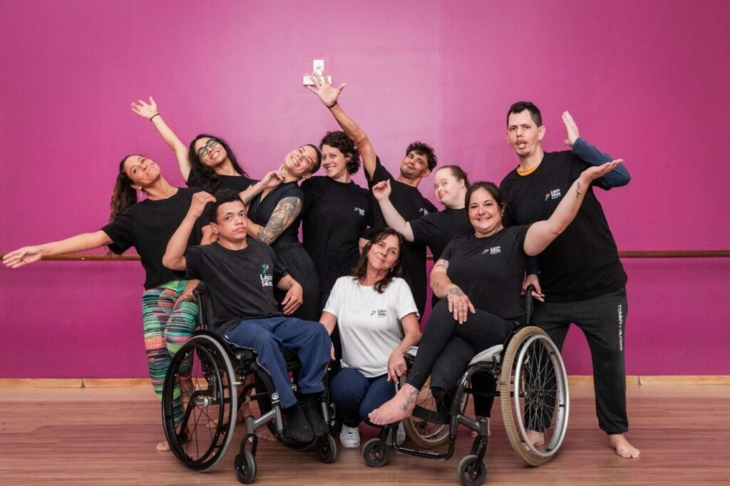 Foto horizontal mostra nove bailarinos, entre homens e mulheres, da Cia Lápis de Seda e a coreógrafa e idealizadora da companhia Ana Luiza Ciscato. Os bailarinos usam camisetas pretas com a logomarca da companhia. Sete deles estão em pé, e fazem diferentes poses para a foto, alguns deles com os braços erguidos. Outros dois estão à frente, sentados em cadeiras de rodas. Ana está agachada no meio deles. Ela é uma mulher de meia-idade, pele clara, cabelos castanhos e lisos na altura dos ombros. Ela usa uma camiseta branca com a logomarca da companhia. Ao fundo, uma parede rosa.